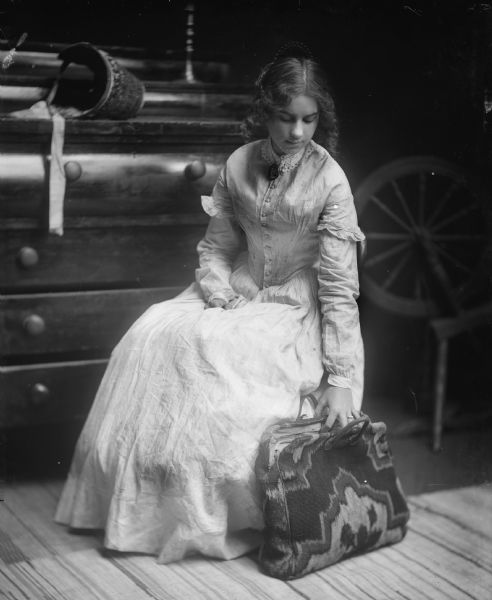 A seated young woman in a nineteenth century style dress poses with a tapestry or carpet bag and spinning wheel.  A bonnet rests on an antiquer dresser behind her.