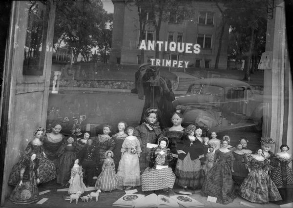 Mr. Trimpey creates a self-portrait while photographing the window of his antique shop at 128 Fourth Avenue. The window display features dolls from the collection of the photographer's wife, Alice Kent Trimpey, and copies of her new book, <i>Becky, My First Love</i>. An automobile and the Sauk County Courthouse are reflected in the window.
