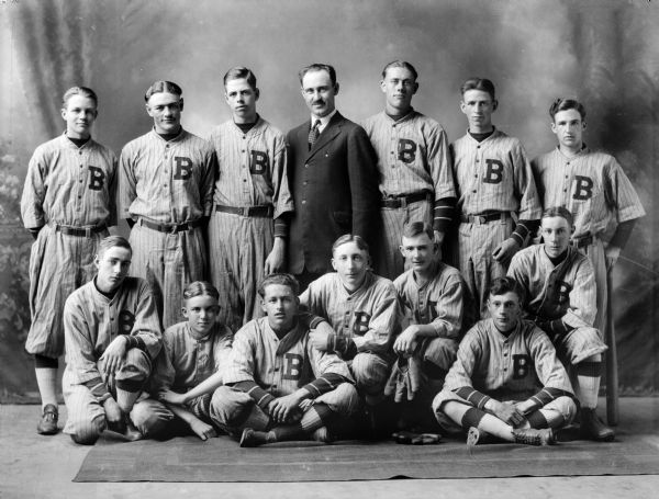 Twelve members of a baseball team, and the bat boy, all in uniform, and one man in a suit, pose for a group portrait. One player holds a mitt, another holds a bat.