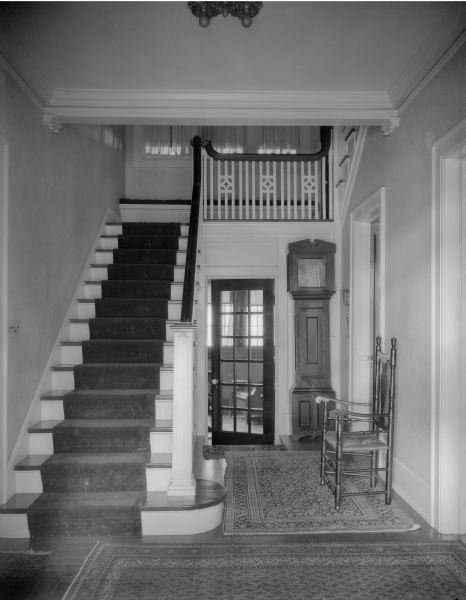 The entrance hall and stairway in the home of William Llewellyn and Zona Gale Breese. There is a grandfather clock and antique rush seat chair in the hall, with oriental carpets on the wood floor.