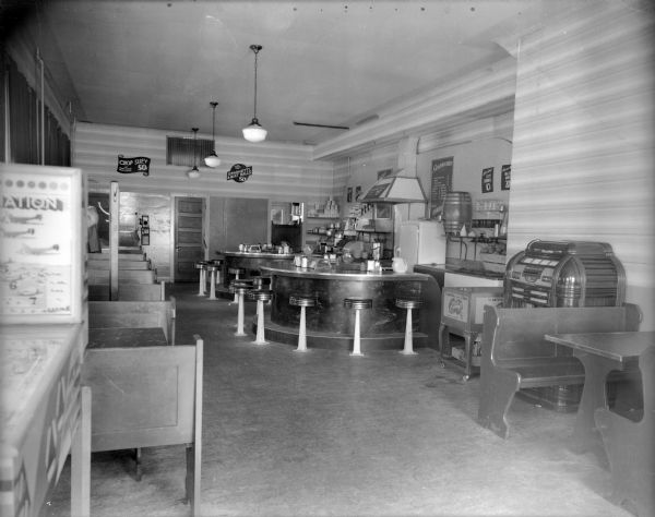 An interior view of a diner, showing two semi-circular counters with stools and booths along the walls. There is a jukebox on the right and a root beer barrel next to the refrigerator. Signs advertise menu items and prices. There are Fiestaware pitchers on the counters. In the foreground on the left is a pinball machine.