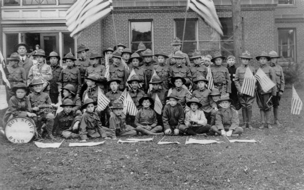 Group portrait of a Boy Scout band in front of a large brick building, possibly the old Baraboo High School. The bass drum head reads "Boy Scout Band" and the boys hold flags and instruments.
