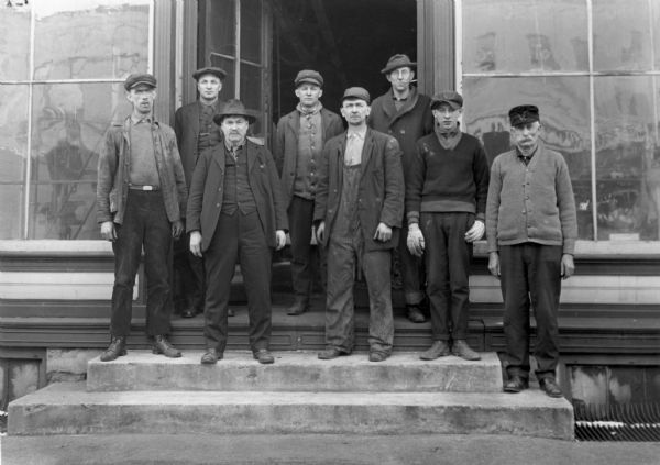 Seven men, five in work clothes and two wearing suits pose on the steps in front of a wooden building, which is a machine shop, with large paned windows. One man, right rear, wears glasses and holds a cigarette holder in his mouth.