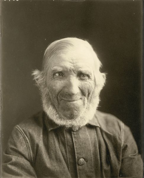 Head and shoulders portrait of an older man with white hair and a neck beard, wearing a denim work jacket. The buttons are embossed with the image of a lantern. On the reverse is written "Mr. Kuball."