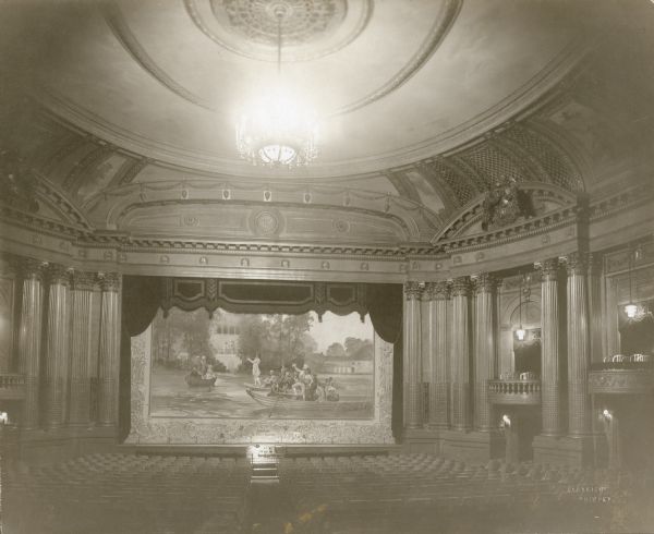 Interior view of the Al. Ringling Theatre highlighting the elaborate woodwork, central chandelier, and stage curtain, which features the painting "Serenade au Petit Trianon." A gooseneck lamp illuminates the organ console; two boxes are seen on the right.    
