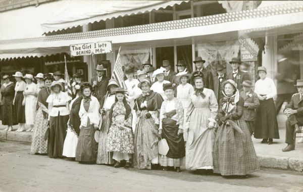Photographic postcard of a group of men and women dressed in clothing of the mid-nineteenth century posing at the side of a Baraboo Street. Most of the women are holding fans. Two women are holding American flags; a man in the background is holding a sign saying: "The Girl I left [sic] Behind Me." Several women on the sidewalk at left are wearing clothing from the time the photograph was taken. A long patriotic banner is hanging above the sidewalk. There are large awnings on the buildings behind the group.