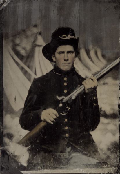 Tintype and hand-colored portrait of John Hodges (who lived in La Fayette), Company I, 28th Wisconsin Infantry, holding his musket. He enlisted on August 21, 1862 and was mustered out of service on August 23, 1865. The background appears to be a painted depiction of a row of tents.