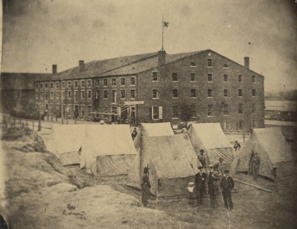 Exterior view of Libby Prison, a Confederate Prison. The view includes tents and three tenement (loft style) buildings. A group of men stand in the foreground near the tents.