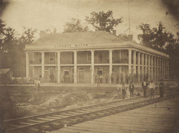View across railroad tracks of men standing in front of and around Osborne House, a large building with columns and a second-story porch.