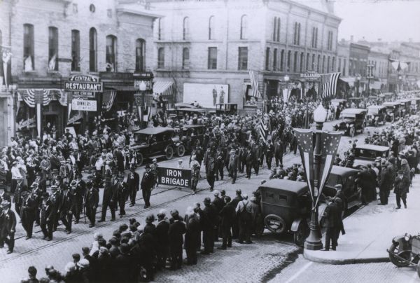 Elevated view of Union veterans, some holding a banner reading "Iron Brigade," marching on a downtown street in a parade during a state encampment.