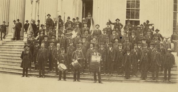 Group portrait of veterans of the 36th Wisconsin Regiment posed on the steps of the Wisconsin State Capital in the late 1870s. Some of the men hold drums and flags.