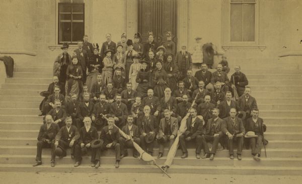 Group portrait of the members of Company F, 16th Wisconsin Volunteer Infantry, and their families, posing on the Wisconsin State Capitol steps during a reunion from September 26-27.

In the front row center is Colonel Reynolds, holding a cane. The second man to his left is James G. Wray. Immediately behind Reynolds is Charles M. Smith with his son Frederick. To Charles Smith's left is J.R. West. On the extreme left of the photo, fourth row back is Dick Adams of Beloit, and the woman to his left is Mrs. Charles Smith. On her left is Will Wray and Glen Wray. Just behind Glen is his sister, Hannah, and to her left is Mrs. Wray.