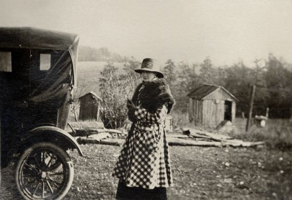 Marjorie Quinney, Richard Quinney's aunt, wearing a stole and a checkered coat, standing behind a Model-T Ford on the family farm with sheds in background.