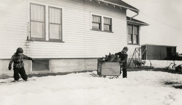 Winter scene with young Richard and Ralph Quinney hauling firewood on a sled through the snow past the farmhouse.
