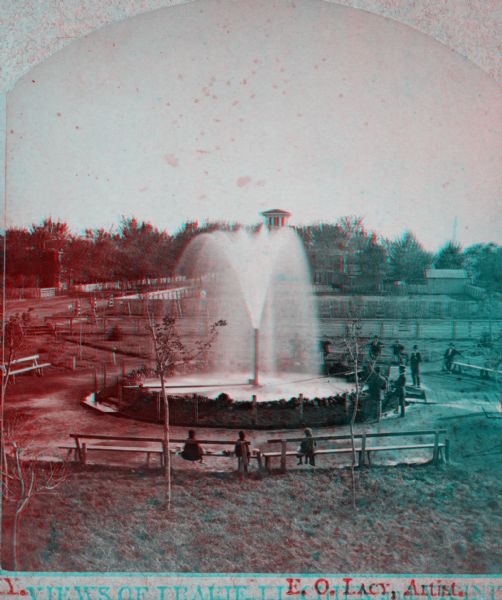 Stereograph of the Artesian Well near the intersection of Wisconsin and Minnesota (later renamed Wacouta) Streets. The following is printed on the back of the photograph:
"The Greatest Artesian Well in America.
The Largest Flow in the World, of the Same Size Bore, with One Exception.
This well was completed in February 1876, bore 5 5/8 inches; pipe projects 10 feet above the ground the water falling into a basin 30 feet in diameter. A movable cap on top of the pipe obstructing the upward flow, and the water is forced outward and upward in all directions, by a pressure of 20,000 pounds.
Depth 960 feet or 970 from top of pipe. Temperature 56 F. Water flowed to top of pipe at a depth of 268 feet. Discharge per minute 603 9-10 gallons or nearly 30,000 bbls. Water will rise in a pipe 60 feet above ground or 100 feet above the Mississippi river. The whole distance that the water will rise is 1,020 feet above the bottom of the well. The pipe goes down 147 feet through sand and gravel to rock, first lime-rock 2 feet, shale 107 feet, sand rock 118 feet, sandy shale 90 feet, red and yellow ocher 6 feet, sandy shale 25 feet, sand-rock 4 feet (here brine begins to flow), sandy shale 71 feet, red, white and yellow sandstone in alternate layers, 355 feet, conglomerate of water-worn quartz-pebbles 35 feet, to bottom of well; which is 327 1/2 feet below the level of the ocean.
The water from this well is found to possess rare medicinal properties, and has cured many diseases of the bladder and kidney, Rheumatism, Dispepsia, St. Vitus dance, chronic female complaints, and has infused new life and vigor into the aged and debilitated."