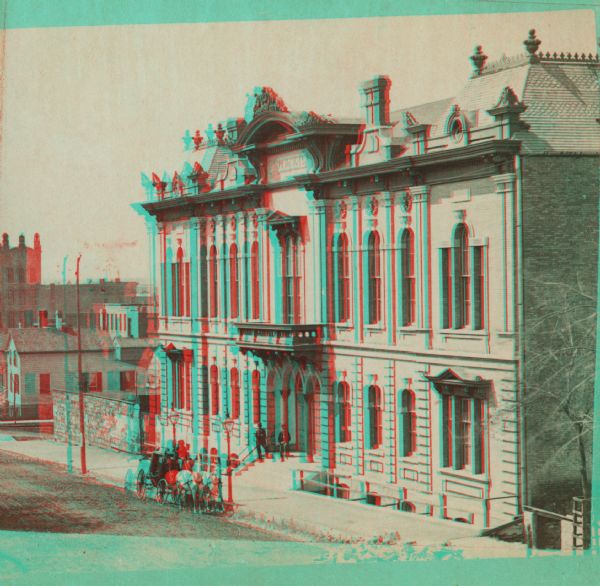 Stereograph; front of music hall along the road, with a horse and carriage team in front of the entrance.  Buildings can also be seen in the left background.