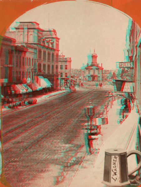 Stereograph; view of the earlier City Hall building, on Market Square at the corner of E. Water Street and Oneida Street. Several signs are in the image, including one in the lower right shaped like a mug with "L. Wagner" on the side. Snow lies on the sidewalk and on the overhangs in front of stores. Two people are standing next to a lamppost in the lower right, and a number of large barrels are on the sidewalk on the left.