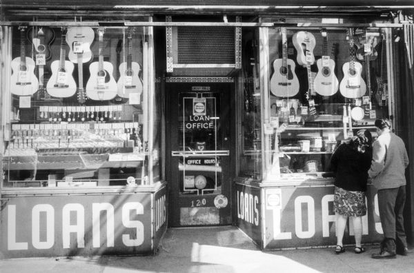 The front of a pawnshop in the East Village showing windows full of merchandise such as guitars and watches above large signs which proclaim, "Loans."  A couple peer into the window on one side of the store.
