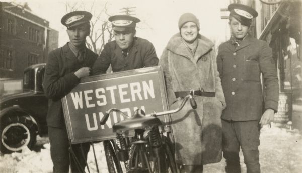 Three Western Union messenger boys with two bicycles and one woman stand on the sidewalk in front of the branch office at 650 State Street. There is snow on the ground and they are leaning on a "WESTERN UNION" sign. The messenger boys were usually university students.