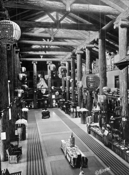 Elevated view of the Glacier Park Hotel lobby. View features the large rustic lobby with log beams and columns, Japanese lanterns, and small gift shops. Chairs and furniture are scattered throughout the lobby.