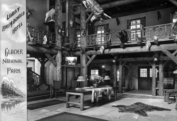 View of the Lake McDonald Hotel lobby. View features the rustic lobby, decorated with animal skins and trophies. There is a balcony overhead, and several chairs and pieces of furniture are arranged throughout the lobby and a set of stairs stands to the far left.