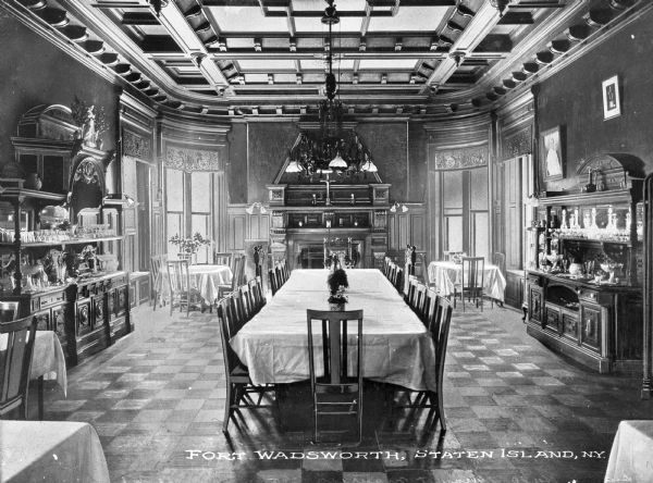 View of an elaborate dining room. View features a long rectangular table surrounded by chairs, two other tables with chairs in the background, a fireplace behind the main table, a decorative ceiling, decorative windows, detailed wood cabinets on each side of the table, and a chandelier hanging above the center table.
