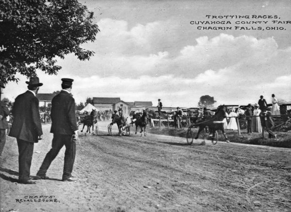 Horses pull wagons on a stretch of racetrack at the Cuyahoga County Fair while spectators gather around the track beside parked automobiles.