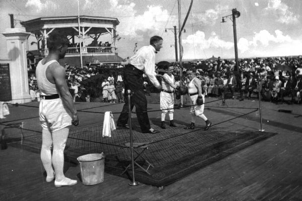 Spectators watch as two dwarfs box in a ring at Midland Beach.  A raised pavilion stands in the background.