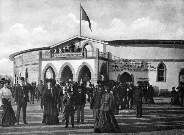 A crowd of men and women in formal attire stands in front of a bullfight stadium. The building features an arched entryway, a balcony, advertisements on the exterior, and a flag on top.
