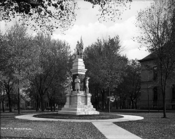 A civil war soldiers' monument stands in on the courthouse ground surrounded by a circular walkway and trees.  Several buildings are visible behind the monument.