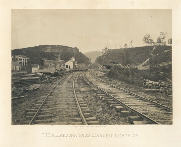 View from railroad tracks through the Allatoona Pass. One of the buildings on the left says "Grocery," and piles of lumber are stacked along the sides of the tracks.<br>Plate 29</br>