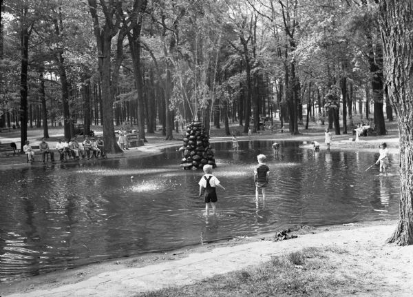 A view of the Marathon County Park. Many children are playing in the water surrounding a stone fountain, while others watch from the benches.