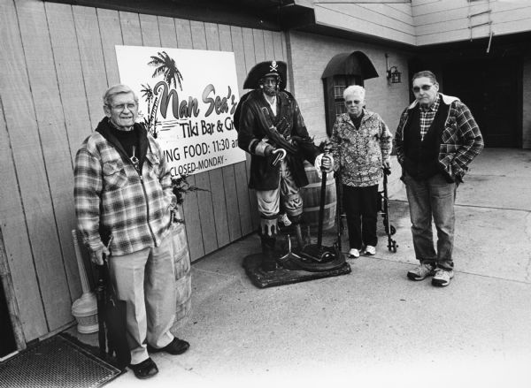"Nan Sea's Tiki Bar and Grill, W921 Kettle Moraine Lane, Dundee (Campbellsport). This was formerly called Frank's On The Lake." From left to right, Ralph "Buddy" Ruecker, John P. Widmer, and Shirley Widmer, behind a statue of a pirate at the exterior of Nan Sea's.