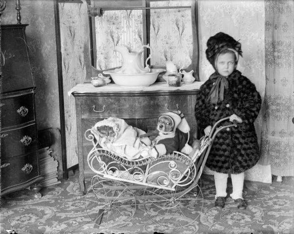 Forest Middleton, dressed in a fur coat and a bonnet, holds the handle of a doll buggy. The buggy carries a dog dressed in a stocking cap and sweater, as well as a cat in bonnet and blanket. In the background, a china commode set sits on a small wooden commode.