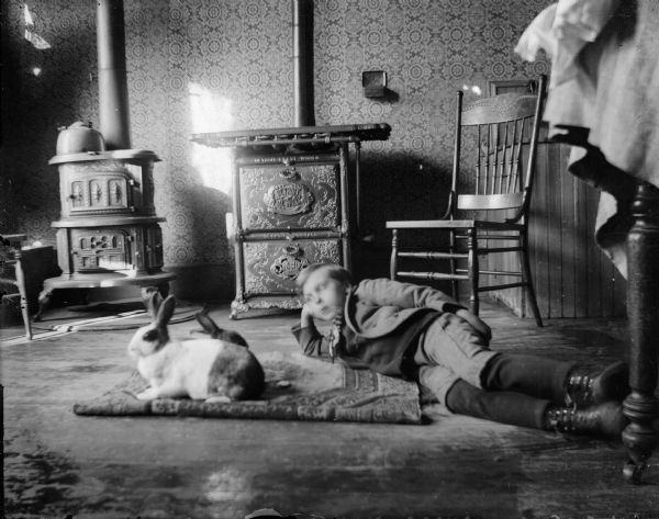 Forest Middleton rests on the kitchen floor with two Dutch hares.  There is a woodburning stove and a "Detroit Jewel" gas stove in the background.  A table leg is visible on the right.