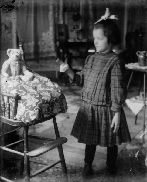 Forest Middleton, wearing a plaid dress, lectures his Teddy Bear, which is seated in a high chair.