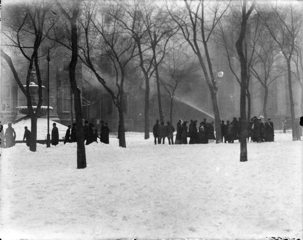 A crowd watches as a stream of water is trained on the south wing of the burning third Wisconsin State Capitol. The Centennial Fountain, with a winter covering over the pool, is on the left.