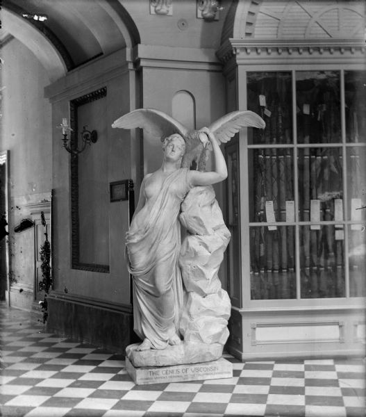 The sculpture, "Genius of Wisconsin" by Helen Farnsworth Mears, displayed in the rotunda of the third Wisconsin State Capitol. On the right is a display case of Wisconsin Civil War flags; a gaslight fixture is on the wall to the left.