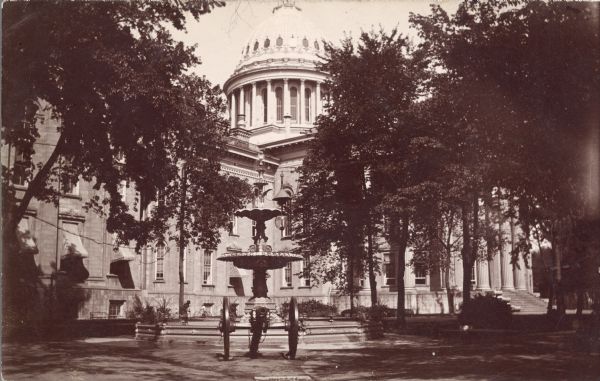 The south side and dome of the third Wisconsin State Capitol, featuring the Centennial Fountain and a cannon. The south wing is to the left; the east wing on the right. There is a boy with a bicycle in the background.