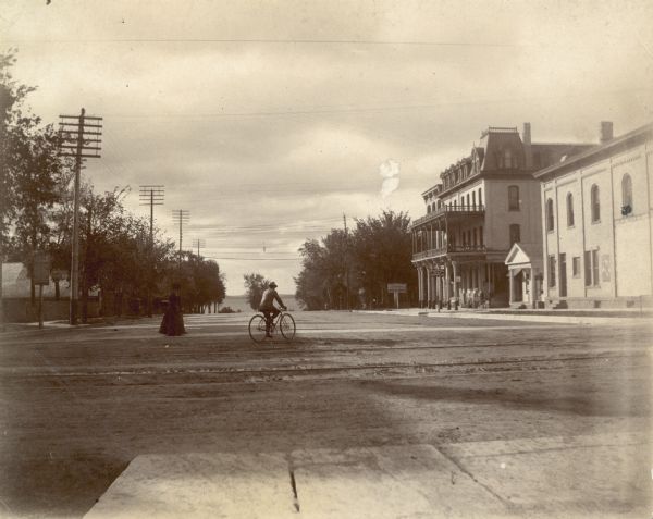 A woman crosses Monona Avenue as a man on a bicycle rides by. The large, Second Empire style Avenue Hotel, with two-story porch, is on the left; the GAR hall is visible beyond it. Lake Monona is in the distance.