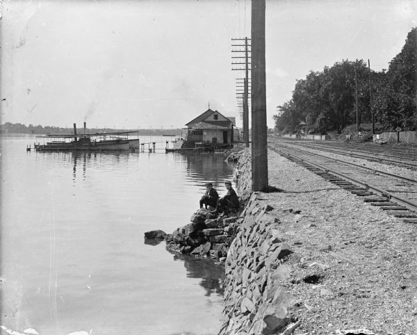 Forest Middleton, right, and a companion sit on the retaining wall along the north shore of Lake Monona near the steamboat dock. Two boats and the boathouse are seen in the background. There are large utility poles and railroad tracks along the shoreline.