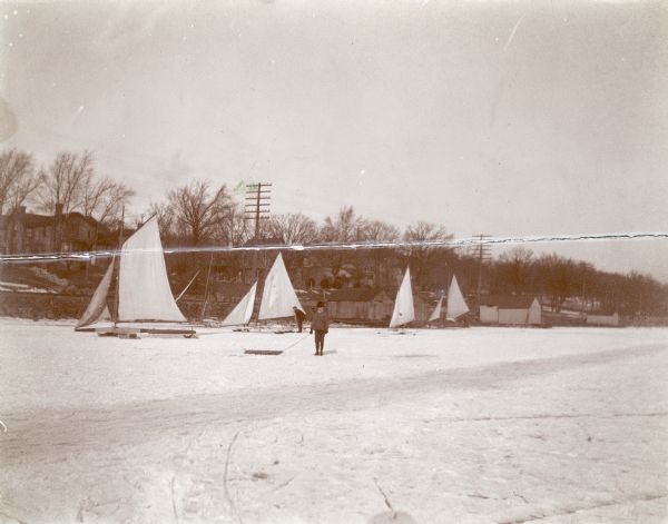 Forest Middleton poses with his sled as his father, William, works with an iceboat behind him on frozen Lake Monona. There are other iceboats and boathouses along the shore.