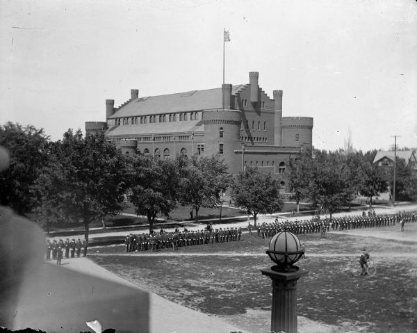 Soldiers stand in formation on the lawn in front of the Historical Society. Bicyclists ride by in the foreground. In the background is the University of Wisconsin-Madison Armory and Gymnasium.