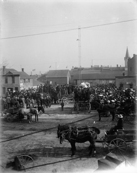 Elevated view of a crowd watching a diver who has launched himself from a high ladder supported by guy wires. The railing around a small pool is visible on the ground. Many of the spectators stand on wagons loaded with wood. There are teams of horses with wagons and buggies in the foreground.