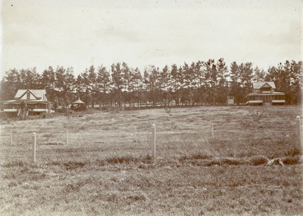 View across fields of two cottages with wrap-around screened porches standing in front of a row of evergreen trees. The house on the right has a second story sleeping porch and outhouse; there is a gazebo near the house on the left. Young trees have been planted in the foreground.