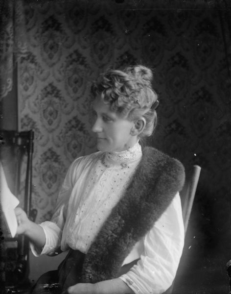 Clara Middleton poses seated in a chair, reading. She is wearing a narrow fur stole over her shoulder.