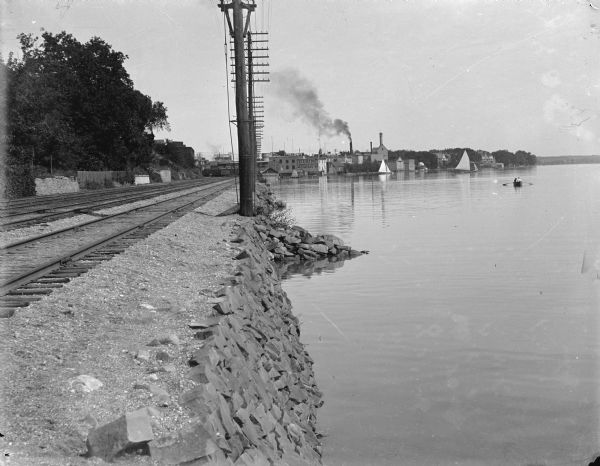 A view east along the north shore of Lake Monona, with Madison's east side industrial area in the background. Tall utility poles and railroad tracks follow the shoreline. The Deering Harvester Company is identified by a sign painted on its wall; large letters on the roof identify the "Advance Thresher Co." The tallest tower right of the smokestack is part of the Fauerbach Brewery complex. The short domed structure on the shoreline is a gazebo which was also a part of the Fauerbach complex. Large private homes are seen on the far right.   There are people in a rowboat and in sailboats in the background.