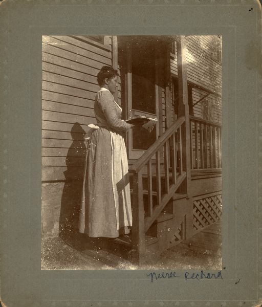 A woman identified as Nurse Rechard stands in profile on the bottom step of a small flight of porch stairs. She is holding a tray with glassware on it.