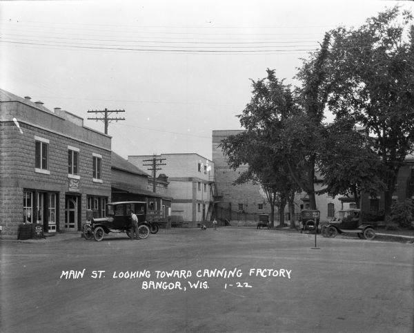 View down street towards a Ford sales and service center on the left, and a canning factory in the background. A man stands near a parked car, and behind him two boys play in the street.