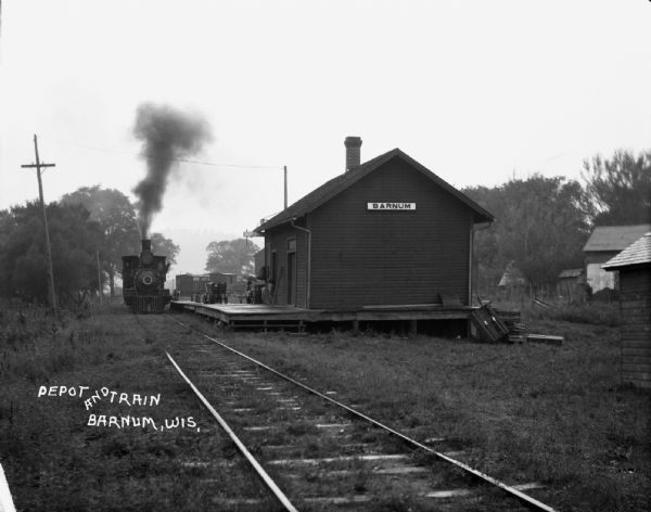 View from along railroad tracks of a train arriving at the depot. There is a child in the doorway of the depot, and a woman in the background is carrying an umbrella.